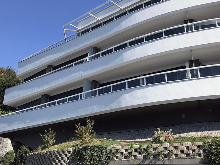 Foto: Gebäude mit hellen Balkonen und Geländer, Detailaufnahme der Obergeschosse, in sich versetze Etagen über der begrünten Natursteinmauer des Kellers