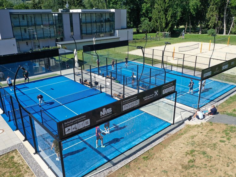Zwei bespielte Padeltennisfelder mit einem Beachvolleyballplatz daneben und einem dreistöckigen Vereinsgebäude dahinter.