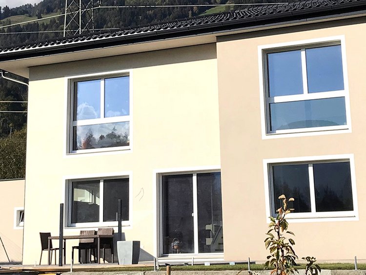 Foto: Frontansicht des Passivhauses, beige Fassade, zwei Fenster im Obergeschoss, zwei Fenster im Untergeschoss, Terrassentüre, im Vordergrund Terrasse mit Steinschlichtung, darauf Tisch und Sessel, Rasen inklusive jungem Obstbaum, im Hintergrund Strommasten, Berg und Wald